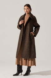A woman is wearing a long, dark brown, plaid-patterned coat with double-breasted buttons and a wide collar. Underneath, she has on a light brown turtleneck sweater and a matching brown skirt that extends below the coat. She is also wearing black high-heeled boots.